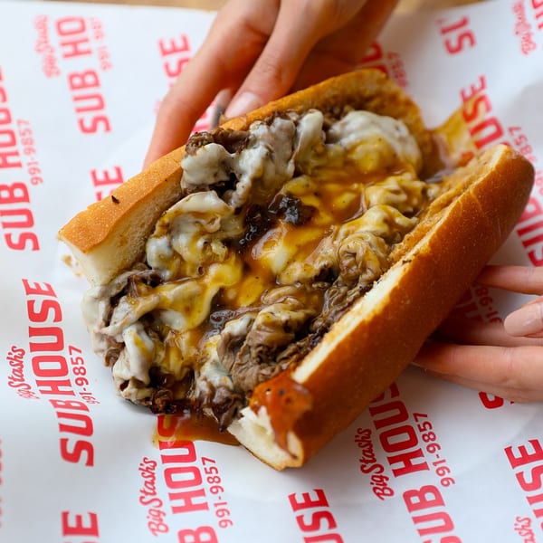 Hands holding a Big Stash's Sub House cheesesteak with melted cheese and gravy on deli paper.