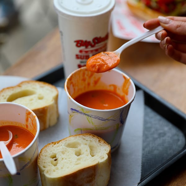 A hand spoons tomato soup from a paper cup, with bread slices and a Big Stash's drink.